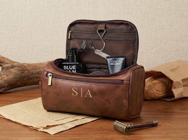 May include: A brown leather toiletry bag with a zipped opening and the initials S|A embossed on the front. The bag is open, revealing an interior with a metal hanging hook, a mesh pocket, and various personal care items inside.