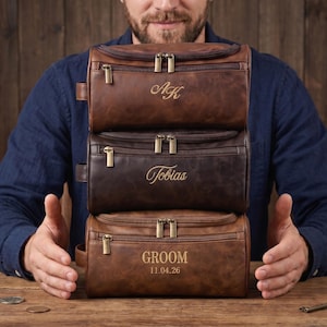 May include: Three stacked rectangular leather toiletry bags in varying shades of brown. The top bag features the initials AK, the middle bag is engraved with the name Tobias, and the bottom bag displays the text GROOM and the date 11.04.26.