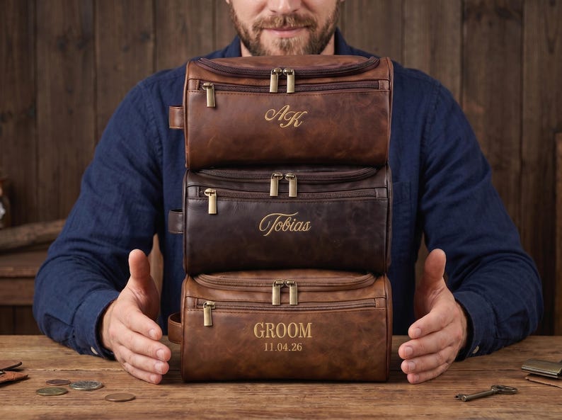 May include: Three stacked rectangular leather toiletry bags in varying shades of brown. The top bag features the initials AK, the middle bag is engraved with the name Tobias, and the bottom bag displays the text GROOM and the date 11.04.26.