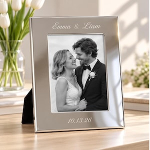 Può includere: Una cornice portafoto rettangolare in metallo color argento con i nomi incisi Emma & Liam in alto e la data 10.13.26 in basso. La cornice contiene una fotografia in bianco e nero di una coppia in abiti formali da matrimonio.