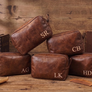 Op de afbeelding: Vijf bruine lederen toilettassen met een gladde, verweerde afwerking. Elke tas is voorzien van een uniek monogram van twee letters op de voorzijde, waaronder SR, CB, AG, LK en HD. De rechthoekige tassen hebben een ritssluiting en een handvat.