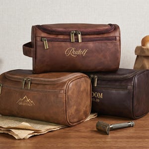 May include: Three rectangular toiletry bags in shades of brown with zippered closures and side handles. The top bag features the name "Rudolf" in script. The front left bag displays a mountain graphic with the name "GEOFFREY". The right bag shows "DOM" and "13.26".