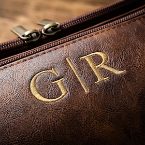 May include: A dark brown textured pouch featuring a gold embroidered monogram with the letters G|R. The pouch includes a dark brown zip closure with two metallic zip pulls at the top edge, showcasing a detailed grain pattern on the surface.