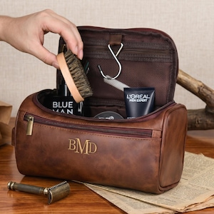 May include: A brown leather wash bag featuring a zipped front compartment and the embossed initials BMD. The bag is open, displaying a dark interior with a metal hanging hook, a wooden brush, and tubes of L'Oreal Men Expert and Blue Man products.