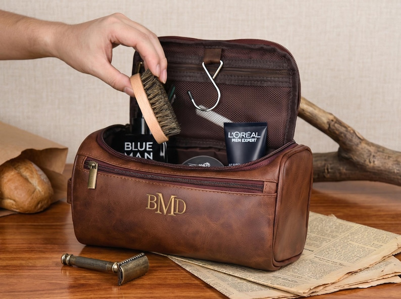 May include: A brown leather wash bag featuring a zipped front compartment and the embossed initials BMD. The bag is open, displaying a dark interior with a metal hanging hook, a wooden brush, and tubes of L'Oreal Men Expert and Blue Man products.