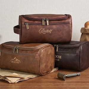 May include: Three rectangular toiletry bags in shades of brown leather. The top bag features the name Rudolf. The front left bag displays a mountain graphic and the name Geoffrey. The front right bag shows the name DOM and the date 13.26.