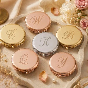 May include: A collection of six round, metallic compact mirrors in gold, silver, and rose gold finishes. Each mirror features a single engraved cursive letter on the lid, including the letters W, C, B, K, Q, and Y, arranged in a circular pattern.