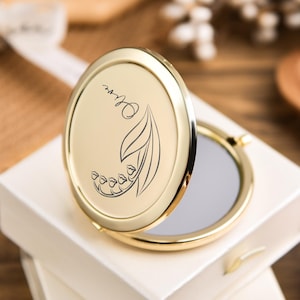 May include: A gold-colored compact mirror with an oval shape, open to reveal a reflective surface. The lid features a floral design with the name "Olive" in script. The compact is resting on a white box, suggesting a gift item.