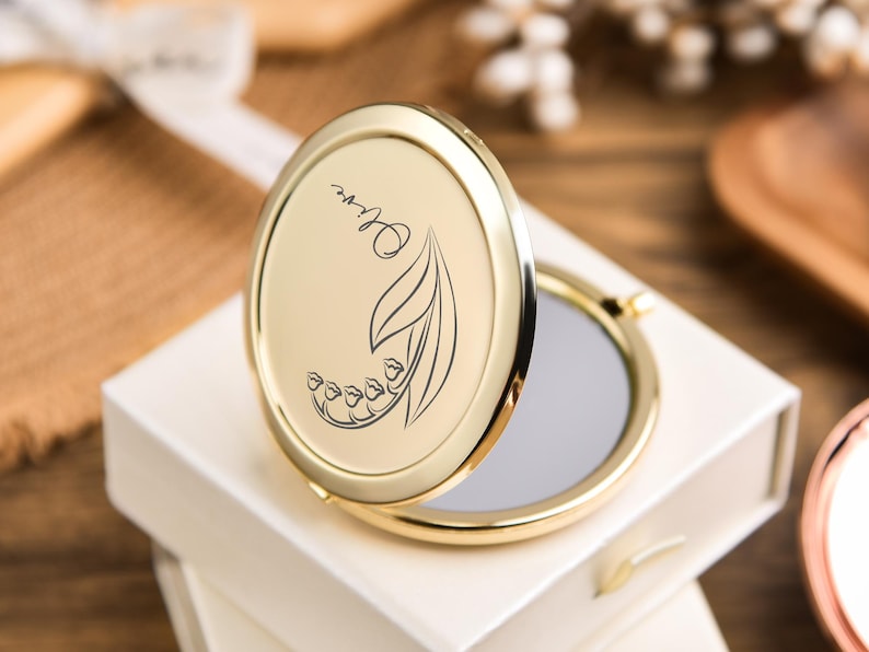 May include: A gold-colored compact mirror with an oval shape, open to reveal a reflective surface. The lid features a floral design with the name "Olive" in script. The compact is resting on a white box, suggesting a gift item.