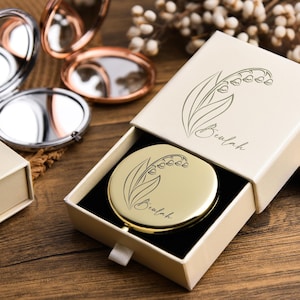May include: A gold compact mirror with a lily of the valley design inside an open cream-colored box. The box and mirror are on a wooden surface, with other compact mirrors in the background.