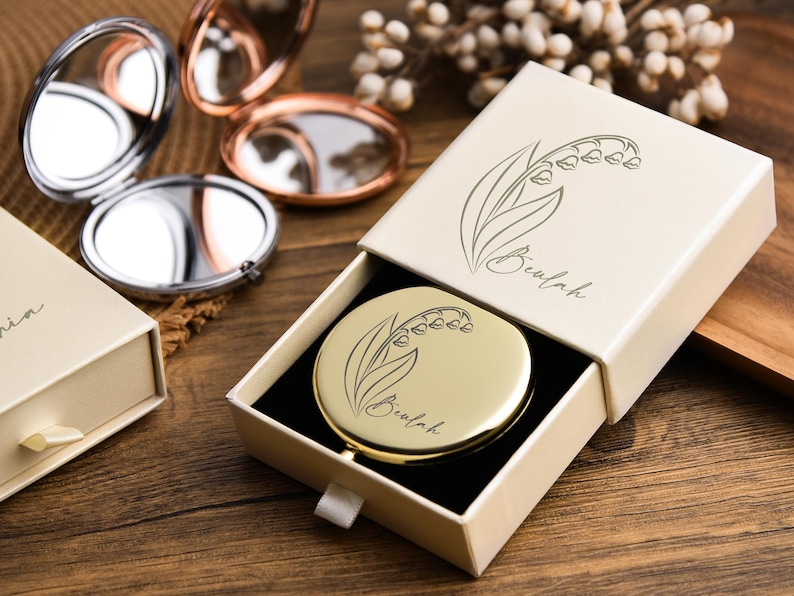 May include: A gold compact mirror with a lily of the valley design inside an open cream-colored box. The box and mirror are on a wooden surface, with other compact mirrors in the background.