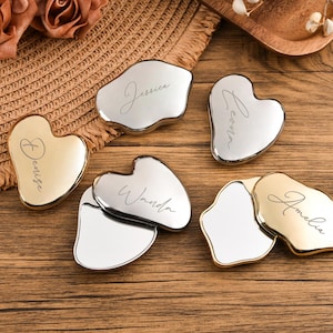 May include: An assortment of personalized gua sha tools in gold and silver tones, each engraved with a different name in cursive script. The tools are in various shapes, including heart and irregular forms, and are arranged on a wooden surface.