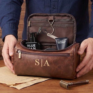 May include: A brown leather wash bag featuring the gold embossed initials S|A on the front. The bag is open, revealing an interior with a hanging hook, a mesh zipped pocket, and various grooming products including a black bottle and a tube.