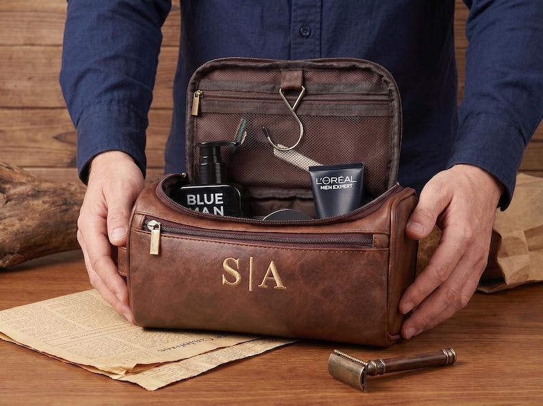May include: A brown leather wash bag featuring the gold embossed initials S|A on the front. The bag is open, revealing an interior with a hanging hook, a mesh zipped pocket, and various grooming products including a black bottle and a tube.