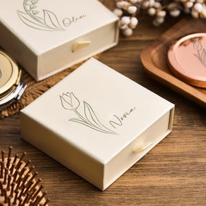May include: Ivory-colored gift boxes and a compact mirror with floral designs. One box features a tulip illustration and the name "Novia." Another box has a lily of the valley design and the name "Olive." A rose gold compact mirror is also visible.