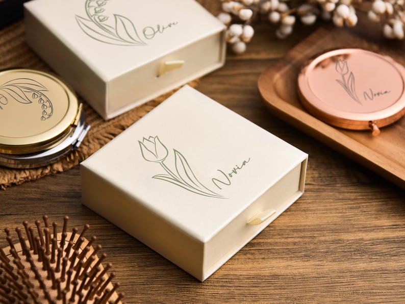 May include: Ivory-colored gift boxes and a compact mirror with floral designs. One box features a tulip illustration and the name "Novia." Another box has a lily of the valley design and the name "Olive." A rose gold compact mirror is also visible.