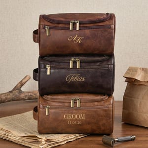 May include: A stack of three rectangular brown leather toiletry bags with brass-colored zippers. The top bag is engraved with AK, the middle bag with Tobias, and the bottom bag with GROOM and the date 11.04.26, each featuring a side handle.
