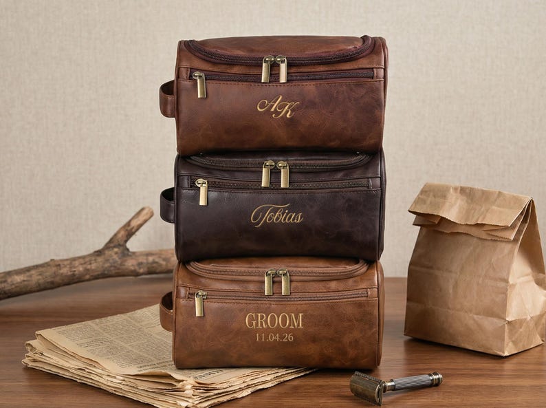 May include: A stack of three rectangular brown leather toiletry bags with brass-colored zippers. The top bag is engraved with AK, the middle bag with Tobias, and the bottom bag with GROOM and the date 11.04.26, each featuring a side handle.