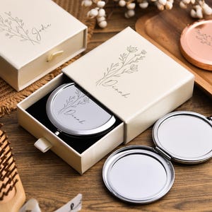 May include: Ivory gift boxes with floral designs and personalized names, one open to reveal a silver compact mirror. A gold and a rose gold compact mirror are also visible, along with a wooden hairbrush.