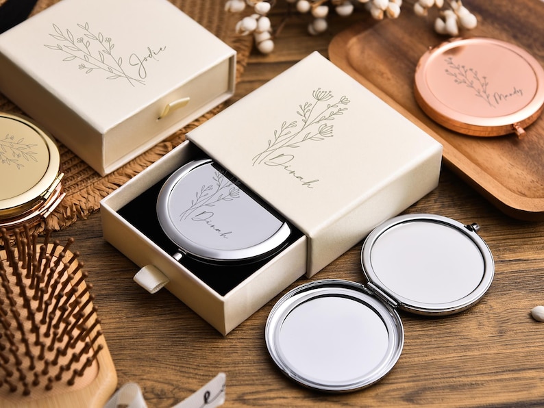 May include: Ivory gift boxes with floral designs and personalized names, one open to reveal a silver compact mirror. A gold and a rose gold compact mirror are also visible, along with a wooden hairbrush.