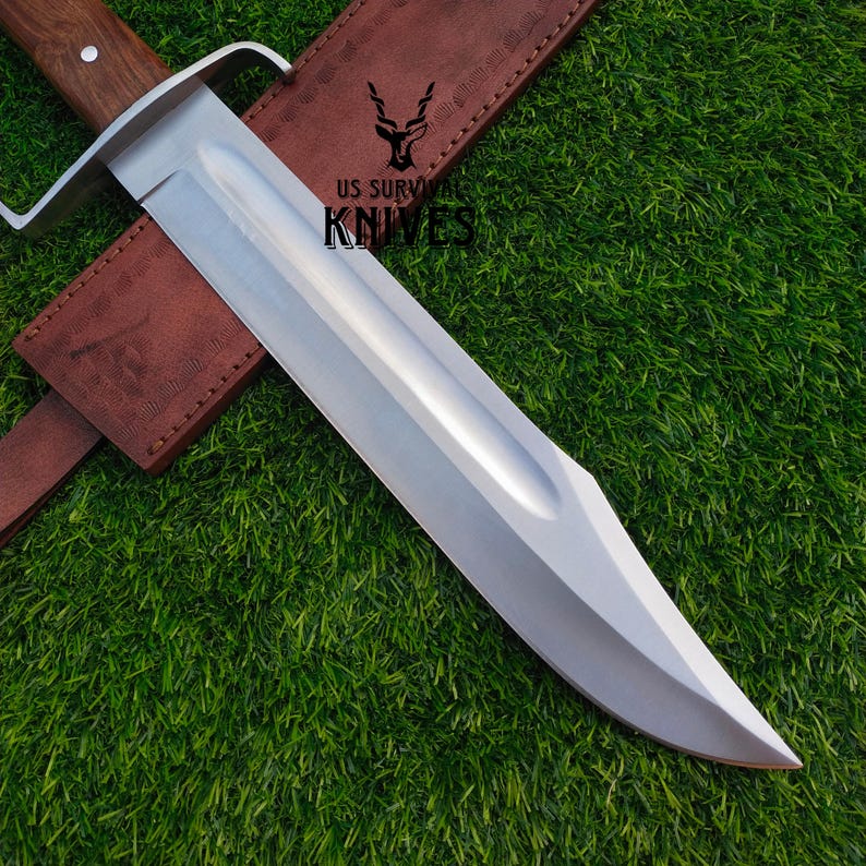 Pode incluir: Uma faca de sobreviv&ecirc;ncia grande, prateada, com cabo de madeira e bainha de couro castanho. A faca tem uma l&acirc;mina larga e uma guarda. O texto "US SURVIVAL KNIVES" &eacute; vis&iacute;vel na bainha.