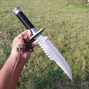 May include: A survival knife with a silver blade and a serrated edge. The handle is black and silver, with a textured grip. The knife has the text "US SURVIVAL KNIVES" and "LURB Support Commando" on the blade.