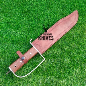 Pode incluir: Uma faca de sobreviv&ecirc;ncia grande com uma bainha de couro castanho e cabo de madeira. A faca tem uma l&acirc;mina larga e uma guarda de metal branco. O texto "US SURVIVAL KNIVES" est&aacute; impresso na bainha.