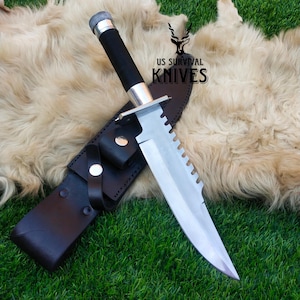 May include: A large survival knife with a serrated blade and a black handle, resting on a leather sheath. The knife has a silver guard and a textured handle. The text "US SURVIVAL KNIVES" is visible.