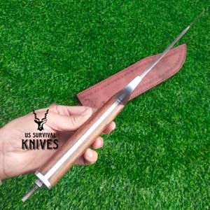 Pode incluir: Uma faca de sobreviv&ecirc;ncia com cabo de madeira e l&acirc;mina de a&ccedil;o inoxid&aacute;vel. A faca &eacute; mostrada com uma bainha de couro marrom. O texto "US SURVIVAL KNIVES" est&aacute; impresso no cabo.