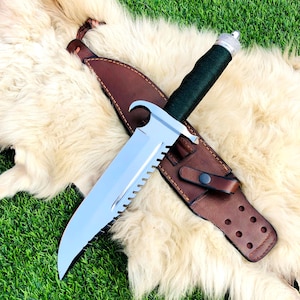 May include: A large, silver-bladed knife with a serrated edge and a dark green wrapped handle. The knife rests in a brown leather sheath with a button closure, set against a backdrop of faux fur and green grass.