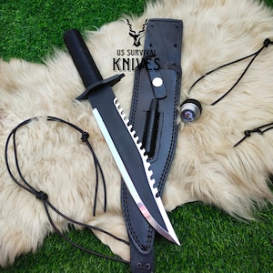 May include: A black survival knife with a serrated blade and a black handle, displayed on a white fur-like surface. The knife is accompanied by a black leather sheath, a compass, and the text "US SURVIVAL KNIVES".