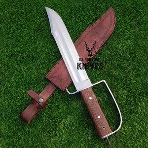 Pode incluir: Uma faca de sobreviv&ecirc;ncia grande com uma l&acirc;mina de a&ccedil;o polido e um cabo de madeira. A faca tem uma prote&ccedil;&atilde;o de dedos distinta e &eacute; combinada com uma bainha de couro marrom. O texto "US Survival Knives" &eacute; vis&iacute;vel.
