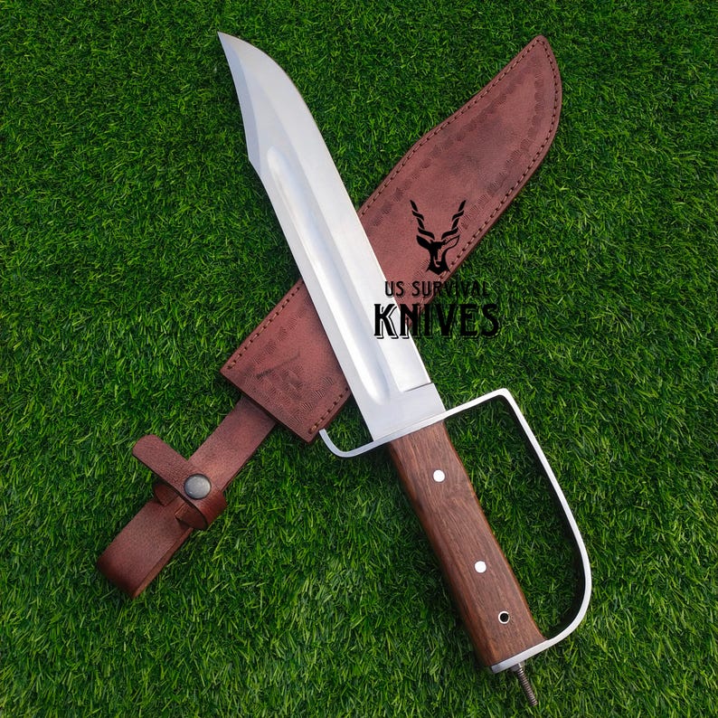 Pode incluir: Uma faca de sobreviv&ecirc;ncia grande com uma l&acirc;mina de a&ccedil;o polido e um cabo de madeira. A faca tem uma prote&ccedil;&atilde;o de dedos distinta e &eacute; combinada com uma bainha de couro marrom. O texto "US Survival Knives" &eacute; vis&iacute;vel.