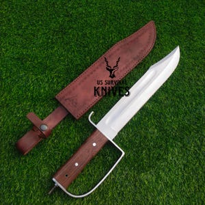 Pode incluir: Uma faca grande de a&ccedil;o polido com cabo de madeira e guarda-m&atilde;o de metal branco. A faca est&aacute; sobre grama verde ao lado de uma bainha de couro marrom. O texto "US SURVIVAL KNIVES" &eacute; vis&iacute;vel.