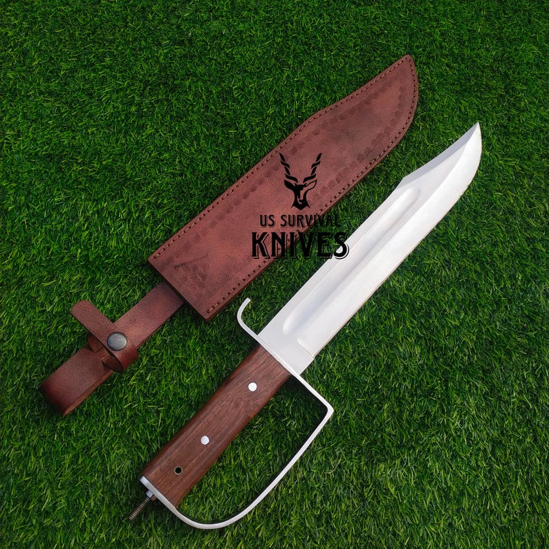 Pode incluir: Uma faca grande de a&ccedil;o polido com cabo de madeira e guarda-m&atilde;o de metal branco. A faca est&aacute; sobre grama verde ao lado de uma bainha de couro marrom. O texto "US SURVIVAL KNIVES" &eacute; vis&iacute;vel.