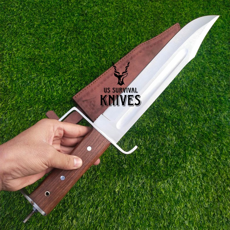 Pode incluir: Uma faca grande de a&ccedil;o inoxid&aacute;vel com cabo de madeira e bainha de couro castanho. A faca tem uma l&acirc;mina larga e uma guarda. O texto "US SURVIVAL KNIVES" est&aacute; impresso na bainha.
