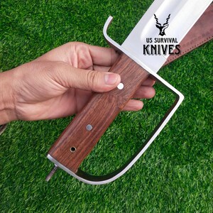 Pode incluir: Uma faca de sobreviv&ecirc;ncia grande com uma l&acirc;mina de a&ccedil;o polido e um cabo de madeira escura. O cabo &eacute; fixado com rebites de metal. A faca tem uma guarda distinta e o texto "US SURVIVAL KNIVES" &eacute; vis&iacute;vel na l&acirc;mina.