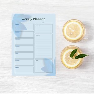 Könnte beinhalten: Ein hellblauer Wochenplaner mit den Worten "Weekly Planner" oben. Der Planer hat Abschnitte für jeden Wochentag. Zwei Gläser Wasser mit Zitronenscheiben befinden sich auf der rechten Seite.