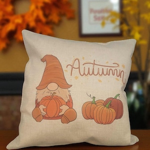 May include: Beige square cushion featuring a gnome holding a pumpkin and the word "Autumn" in orange script. Several orange pumpkins are to the right of the gnome. The cushion is on a wooden surface.