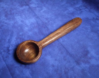 May include: A handcrafted wooden coffee scoop featuring a deep, rounded bowl and a long, smooth handle. The wood displays a rich, dark brown color with visible natural grain patterns throughout the entire surface of the utensil.