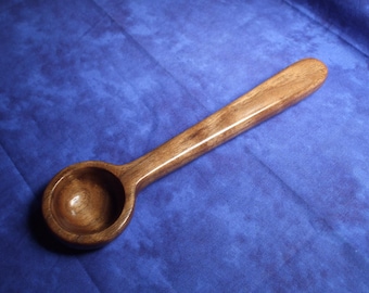 May include: A handcrafted wooden measuring scoop featuring a smooth, polished finish with visible natural grain patterns. The utensil has a deep, circular bowl and a long, straight handle, showcasing the rich brown tones of the wood.