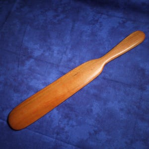 May include: A smooth, light brown wooden paddle with a long, tapered handle and a wide, flat blade. The wood grain is visible across the surface, showing natural variations in color and texture. The object has a rounded tip and edges.