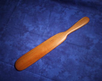 May include: A smooth, light brown wooden paddle with a long, tapered handle and a wide, flat blade. The wood grain is visible across the surface, showing natural variations in color and texture. The object has a rounded tip and edges.