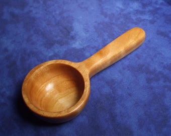 May include: A light brown wooden measuring scoop with a smooth, polished finish. The scoop features a deep, rounded bowl and a thick, sturdy handle extending from the side, showcasing natural wood grain patterns throughout the entire piece.
