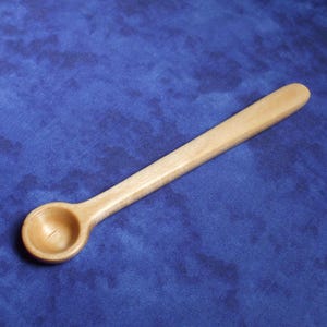 May include: A small, wooden spoon with a rounded bowl and a long, slender handle. The spoon is light brown in color and appears to be handmade. The spoon is set against a blue background.