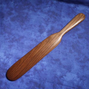 May include: A handcrafted wooden spatula with a long, flat blade and a rounded handle. The spatula is made of dark brown wood and has a smooth, polished finish. The background is a blue fabric.