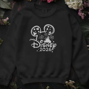 May include: Black crew neck sweatshirt with a white Disney design. The design includes Mickey Mouse ears, fireworks, a castle, and the text "Disney 2026". The sweatshirt is surrounded by flowers.