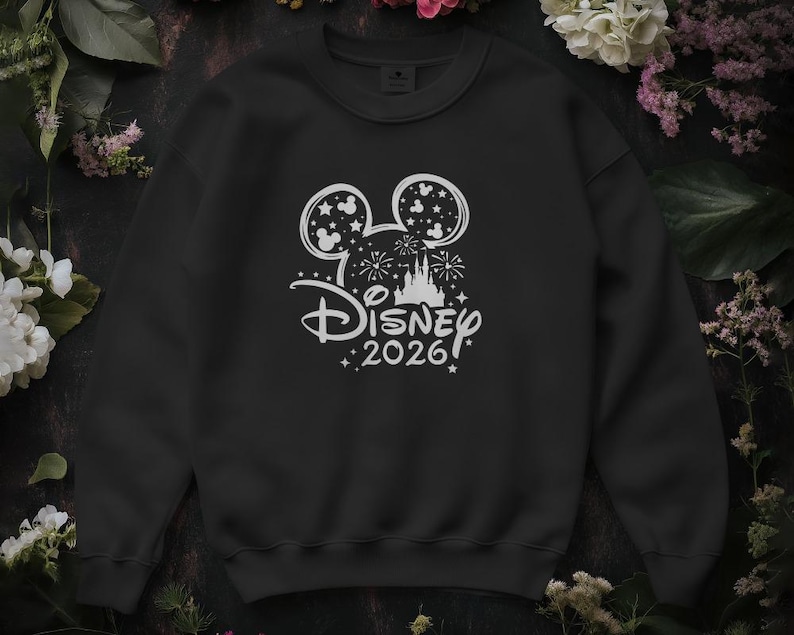 May include: Black crew neck sweatshirt with a white Disney design. The design includes Mickey Mouse ears, fireworks, a castle, and the text "Disney 2026". The sweatshirt is surrounded by flowers.