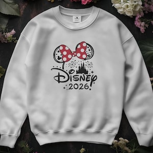 May include: White sweatshirt featuring a Minnie Mouse bow with red polka dots. The design includes the Disney logo, a castle silhouette, fireworks, and the text "2026" in black. A crew neck sweatshirt.