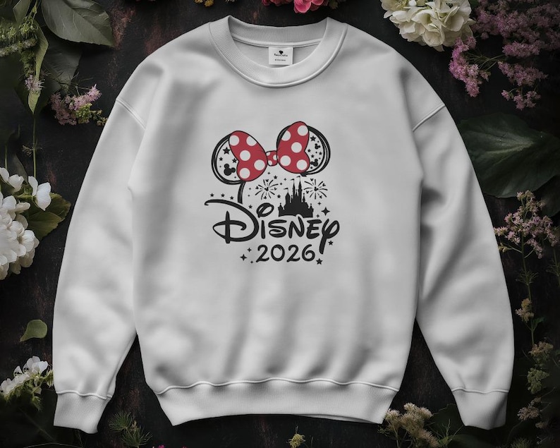 May include: White sweatshirt featuring a Minnie Mouse bow with red polka dots. The design includes the Disney logo, a castle silhouette, fireworks, and the text "2026" in black. A crew neck sweatshirt.
